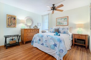 A bedroom with a bed, wooden dresser, two nightstands, a ceiling fan, a window with curtains, and a wall with a framed picture and a poster. - Maui Vista 2310 - Fully Remodeled 1 BR/1 BA Close to Beach (Kihei)