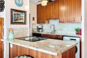 A kitchen with wooden cabinets, a granite countertop, a coffee maker, and a dishwasher. - Maui Vista 2310 - Fully Remodeled 1 BR/1 BA Close to Beach (Kihei)