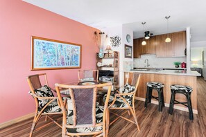 A dining area with a glass-top table and wicker chairs, a kitchen with wooden cabinets and pendant lights, and a pink wall with a framed artwork. - Maui Vista 2310 - Fully Remodeled 1 BR/1 BA Close to Beach (Kihei)