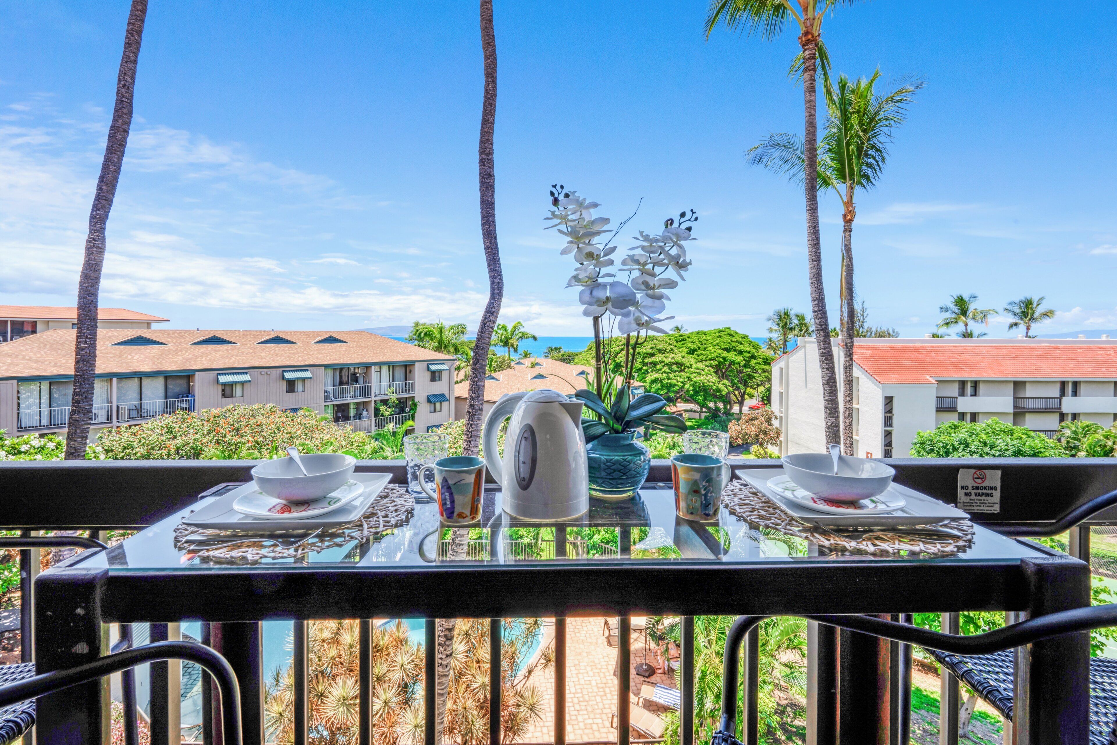 Mesa de comedor dispuesta con platos, tazones y una tetera, con vistas a palmeras y edificios.