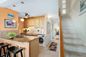 A modern kitchen with granite countertops, wooden cabinets, a built-in washing machine, and a dining area with bar stools. - Maui Vista 2408 - Deluxe 2 Bedroom with Ocean View (Kihei)
