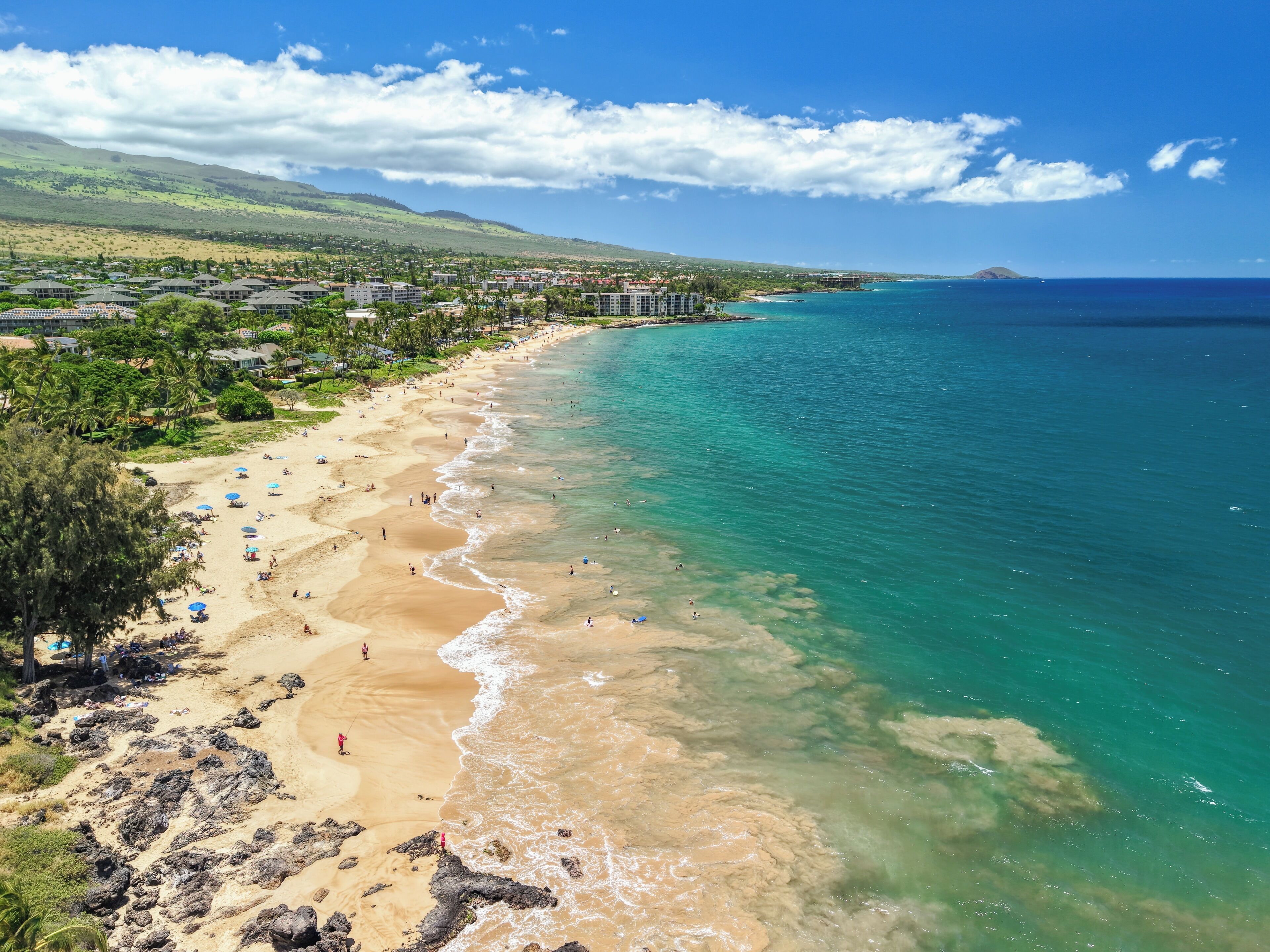 Una playa con agua turquesa cristalina, una orilla arenosa y colinas y edificios al fondo.