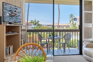 A room with a television displaying dolphins, a bookshelf, a dining area with chairs and a table, and a view of palm trees and buildings outside. - Maui Vista 3212 - Inviting 1 BR/1 BA near Beach (Kihei)