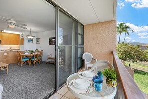 A balcony with a dining table set for four, a sliding glass door leading to a kitchen, and a view of a tropical landscape. - Maui Vista 3212 - Inviting 1 BR/1 BA near Beach (Kihei)