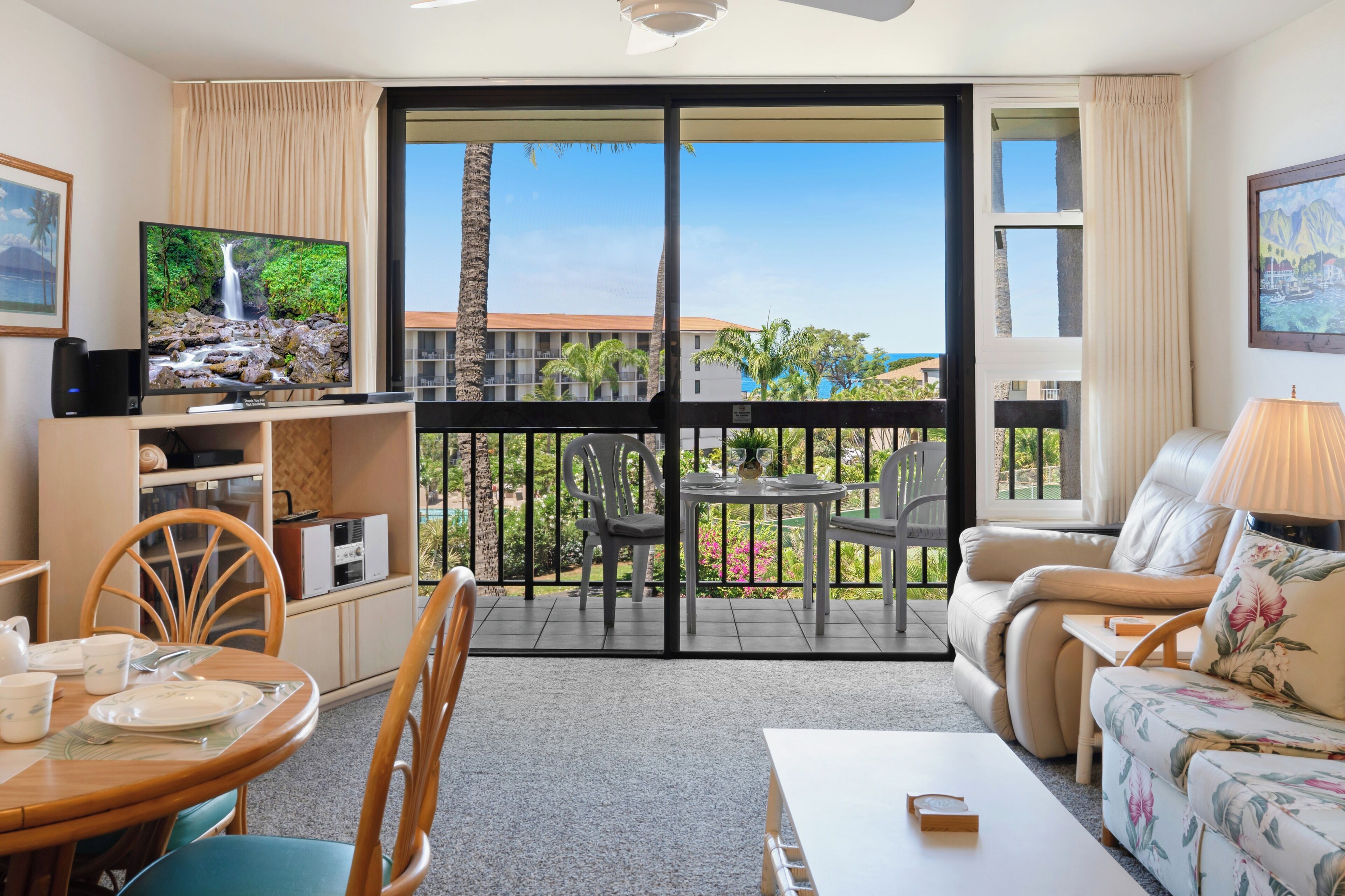 Ein Wohnzimmer mit Fernseher, einem Essbereich und einem Balkon mit Blick auf Gebäude und Palmen.