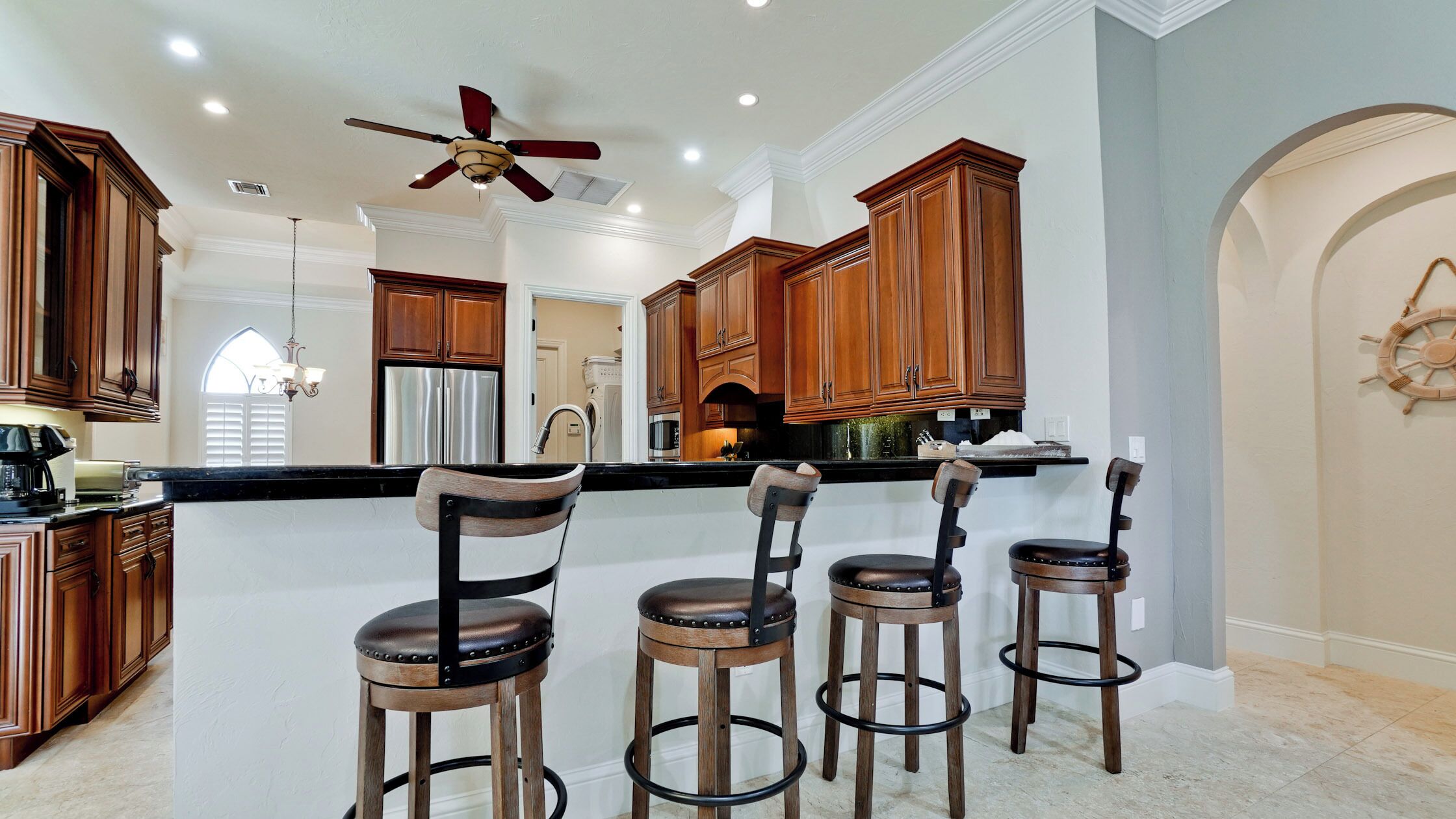 A kitchen with wooden cabinets, a central island with bar stools, a ceiling fan, and a stainless steel refrigerator.