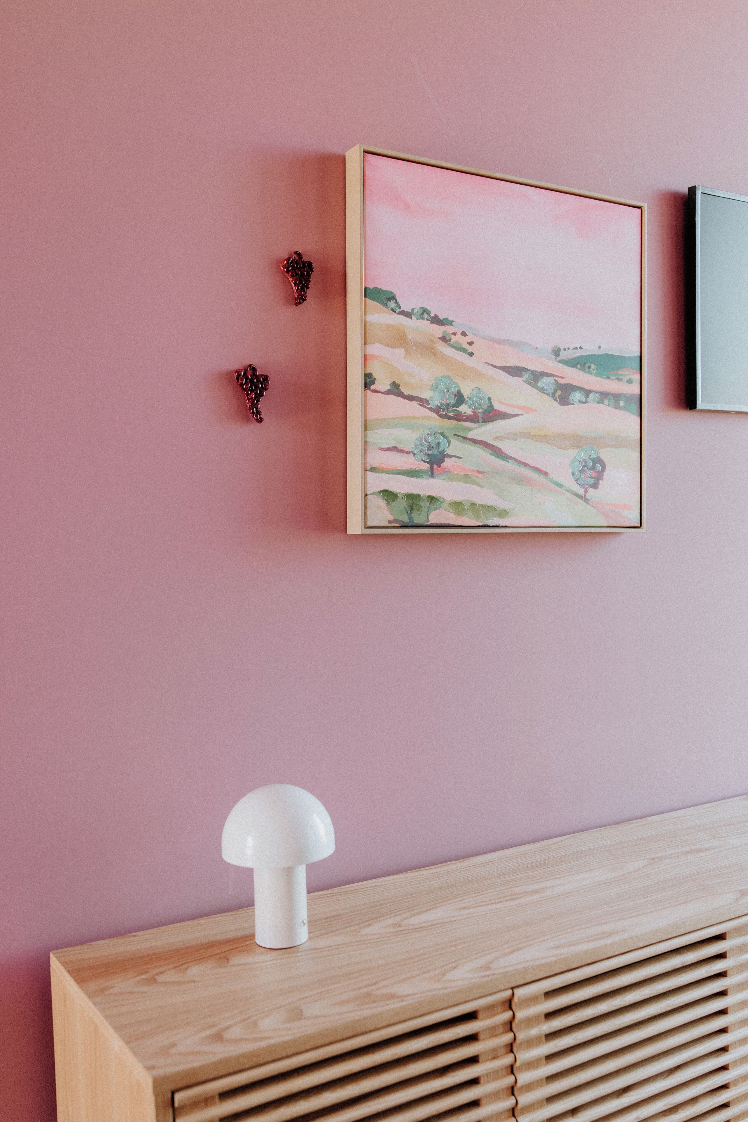 Una habitación con una pared rosa, un cuadro enmarco de un paisaje, una lámpara blanca sobre una consola de madera y un pequeño objeto decorativo.