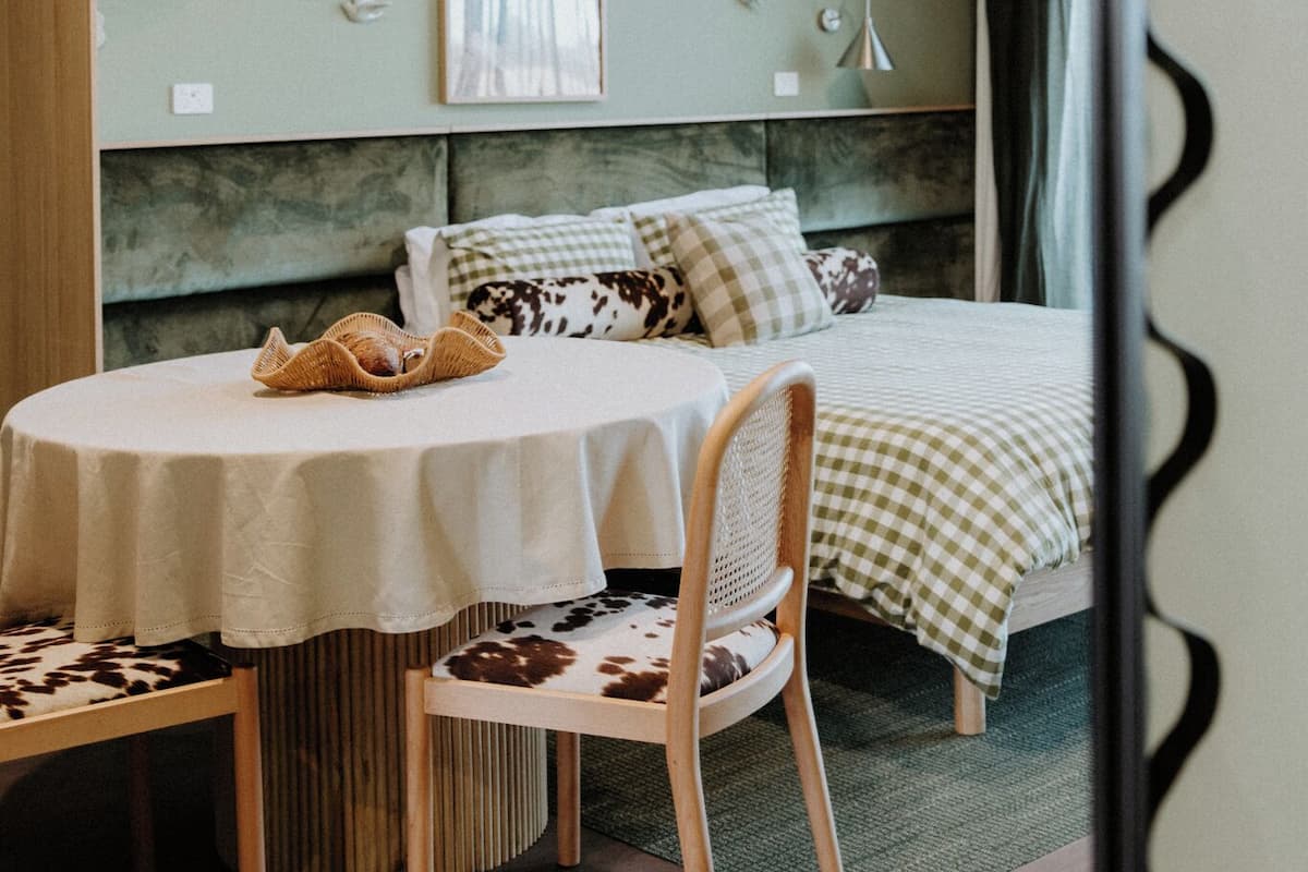 A room with a bed, a round table with a chair, a hanging light fixture, and a wall-mounted artwork.