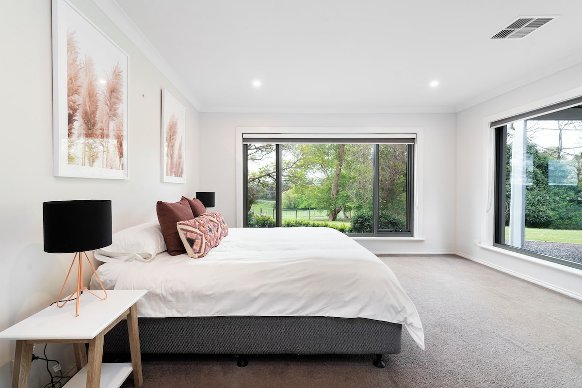 A modern bedroom with a large bed, a bedside table with a lamp, and a view of greenery through large windows.