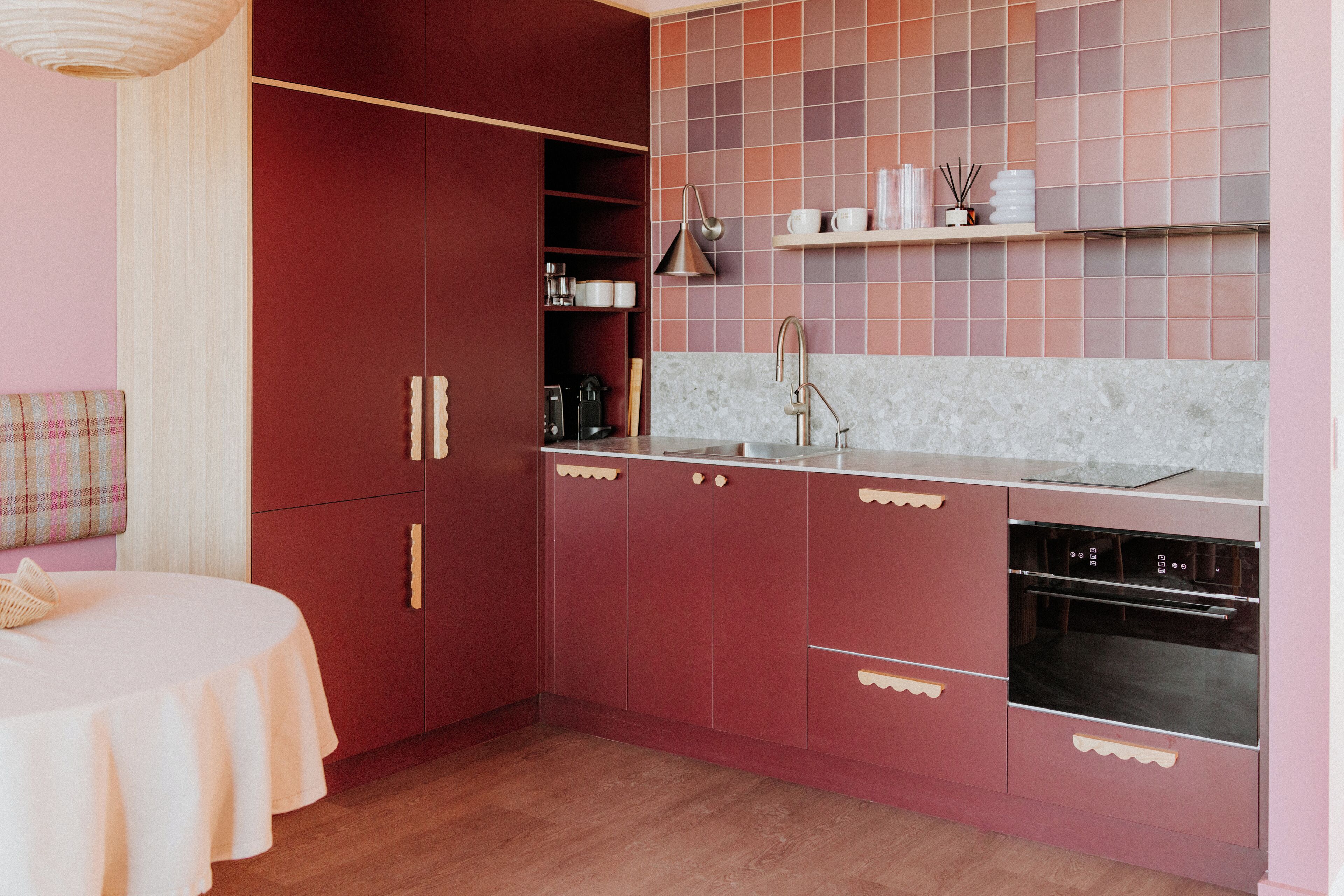 A compact kitchen with a built-in oven, a sink, and a countertop. There is a dining table with a white tablecloth and a plaid cushion.