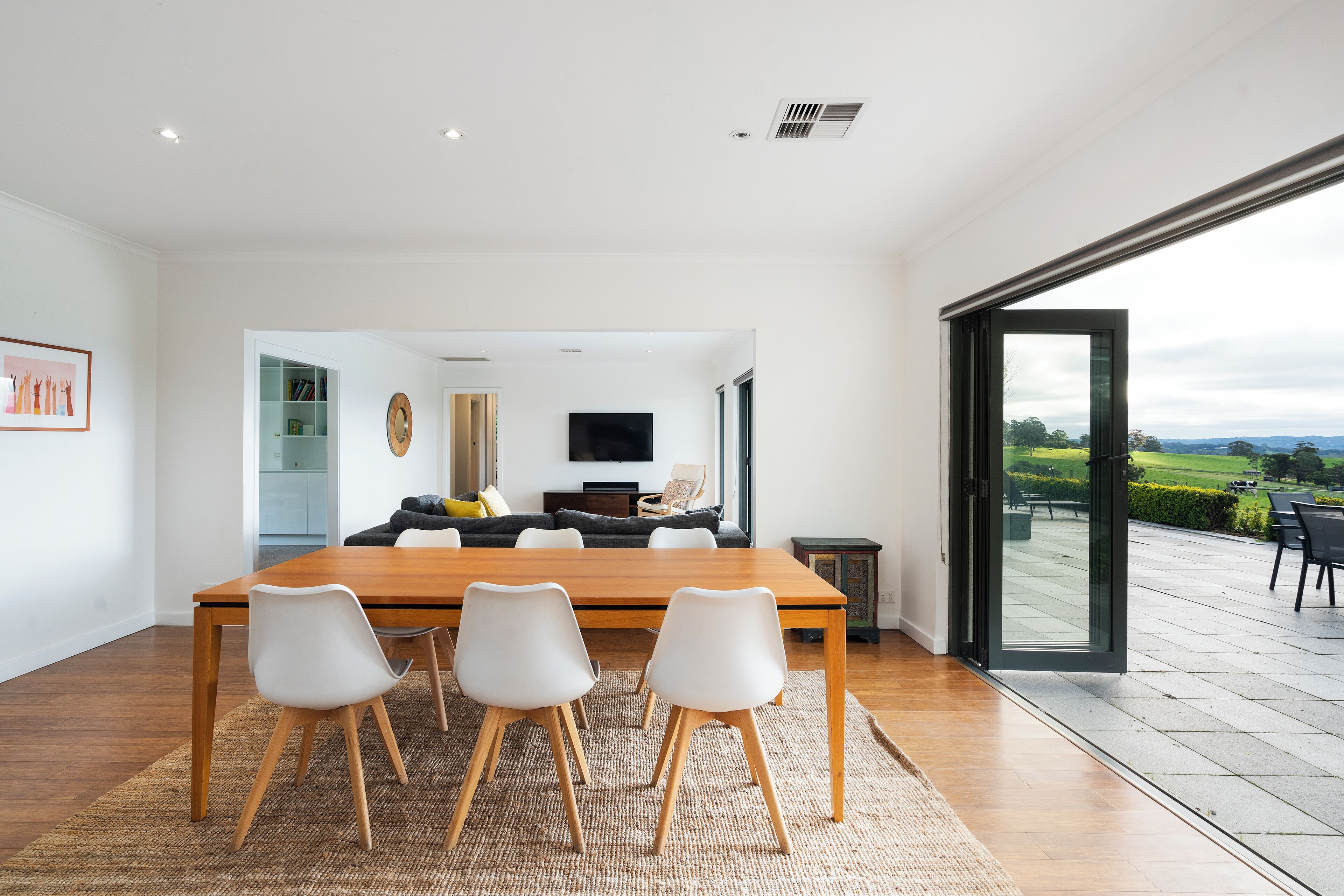 A modern dining area with a wooden table and white chairs, a flat-screen TV, and a view of the outdoors through a glass door.