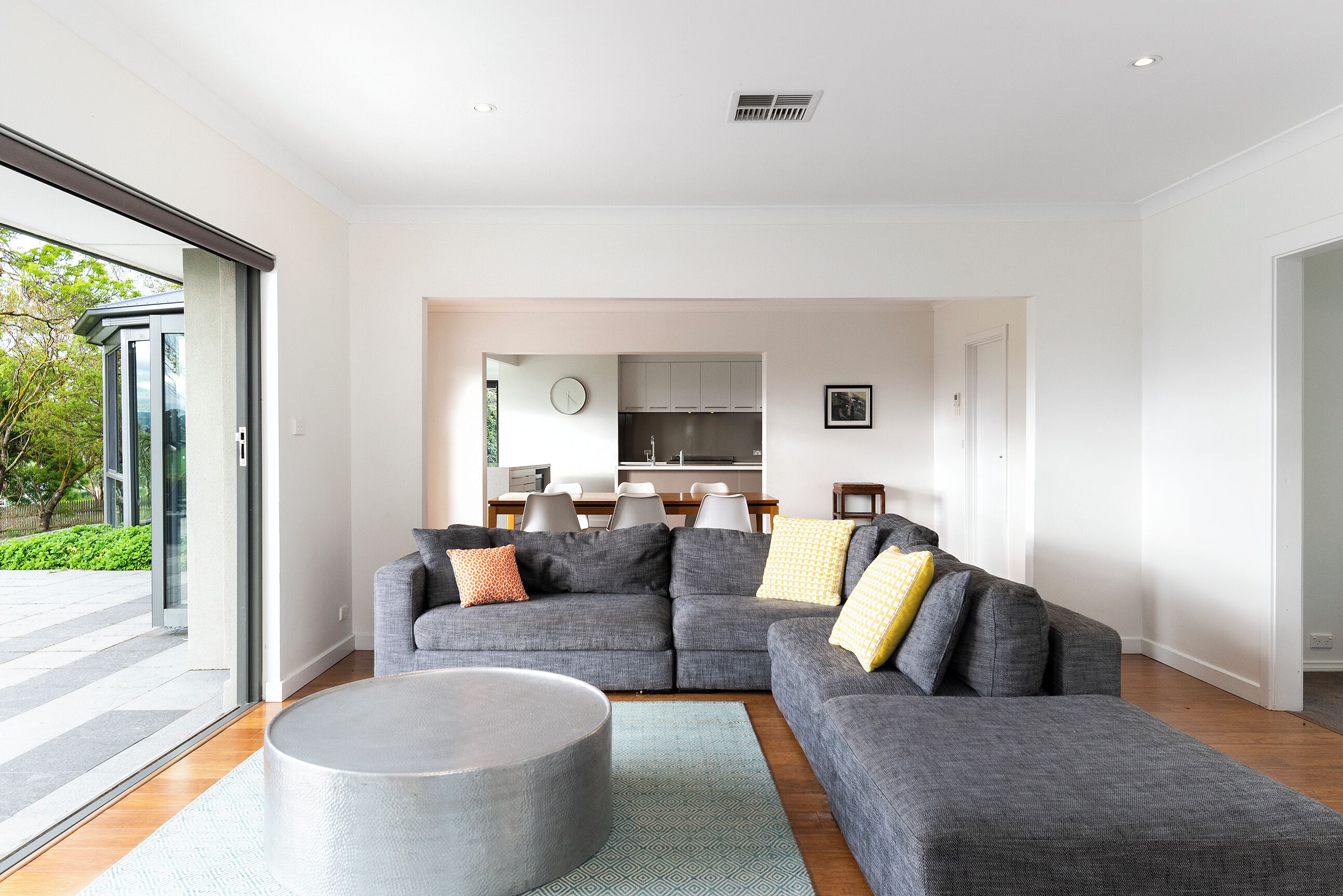 Un salon moderne avec un canapé d’angle, une table basse ronde et une vue sur la cuisine et la salle à manger, grâce à une porte ouverte.