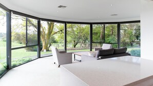 A modern living room with a large white table, two grey sofas, and a curved glass wall offering a view of a garden and trees. - Hahndorf Farm Barn (Hahndorf)