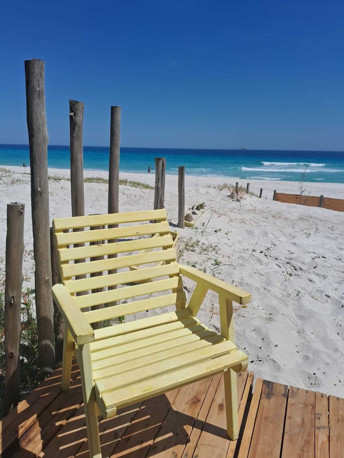 Une terrasse en bois avec un banc jaune donnant sur une plage où l’eau est d’un bleu limpide et le rivage est sablonneux.