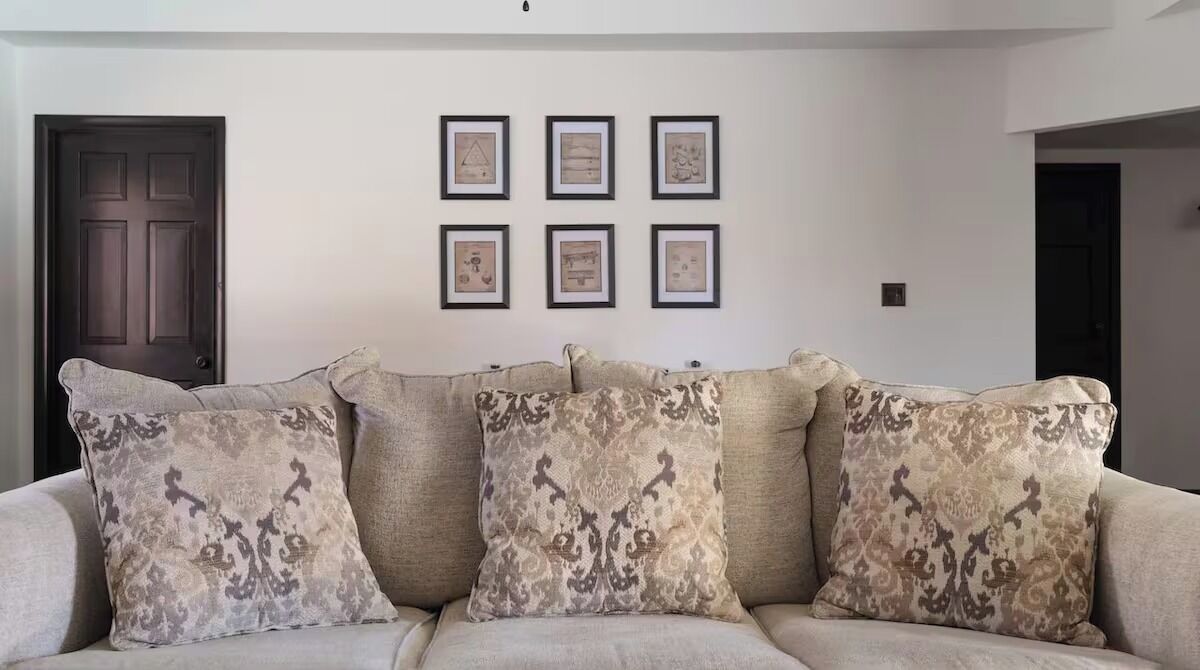 Een woonkamer met een beige bank en gestreepte kussens, een donkerhouten deur en zes ingelijste foto's aan de muur.