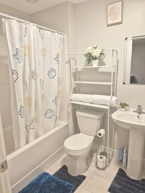 Een badkamer met toilet, wastafel, douchegordijn met schelpenprint, handdoekhouder en een kleine potplant.