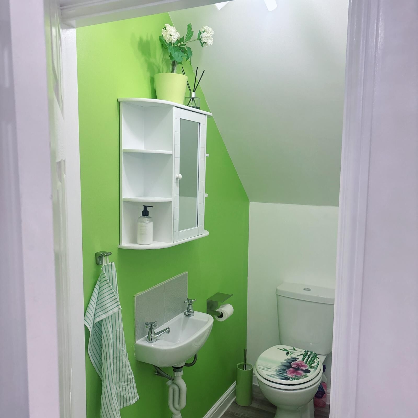 Baño con pared verde, lavamanos blanco, espejo, mueble blanco, inodoro y toalla colgada en la pared.