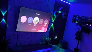 Un salon moderne avec un téléviseur à écran plat fixé au mur, un coin salon avec un pouf blanc et une plante en pot.