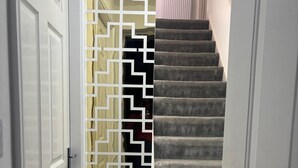 Un couloir avec une porte blanche, un panneau décoratif aux motifs géométriques blancs et dorés, et un escalier recouvert de moquette avec un radiateur fixé au mur.