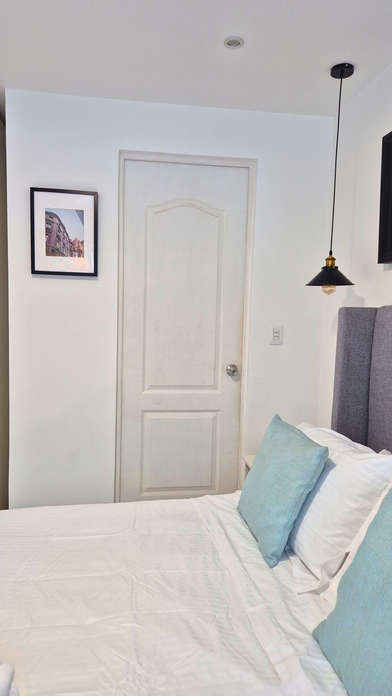 Une chambre à coucher avec un lit, une porte, un tableau encadré, une suspension et une prise de courant au mur.
