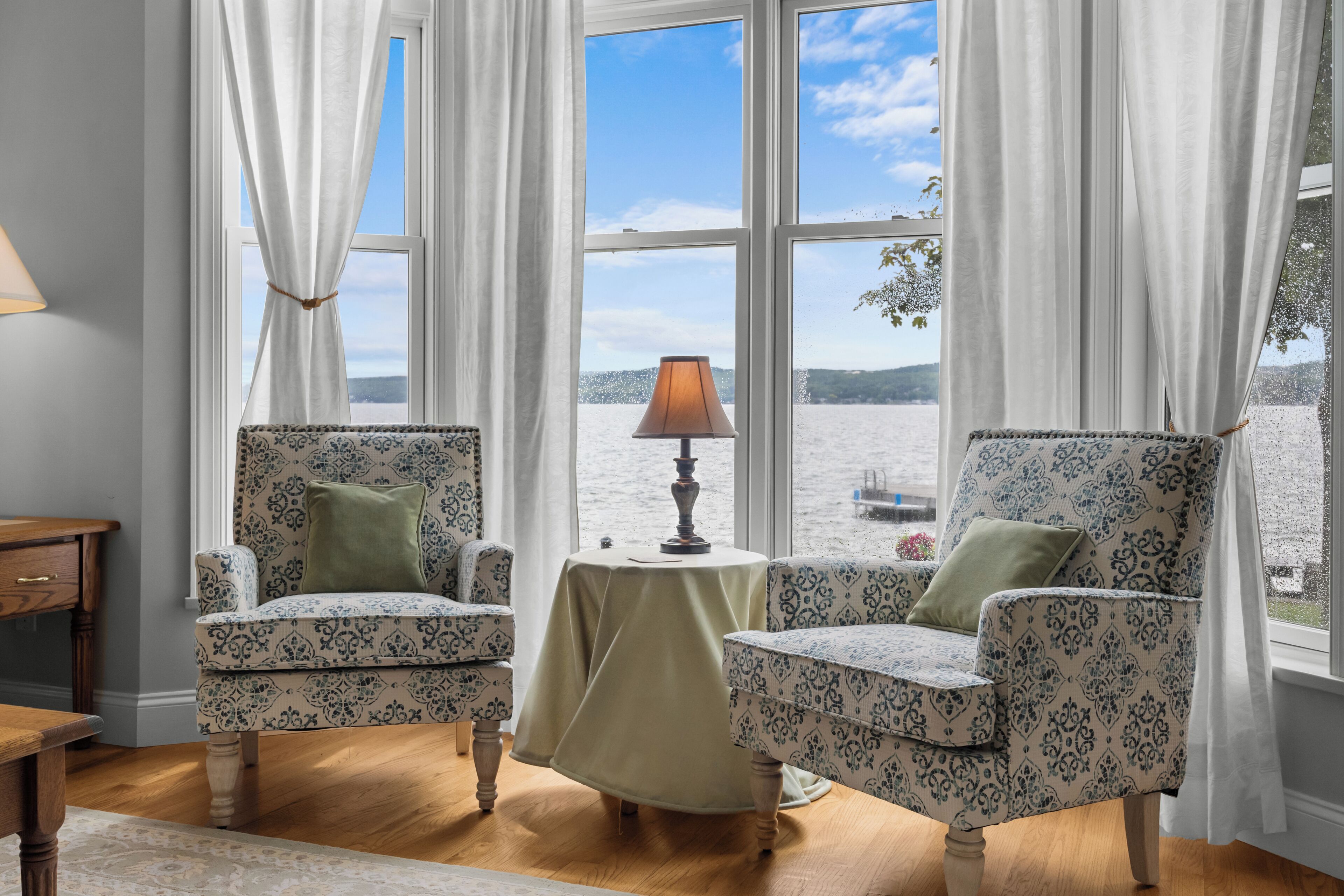 Une pièce avec deux fauteuils à motifs, une petite table avec une lampe et une vue sur une étendue d’eau grâce à de grandes fenêtres.
