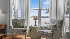 Ein Zimmer mit zwei gemusterten Sesseln, einem kleinen Tisch mit Lampe und Blick auf ein Gewässer durch große Fenster.