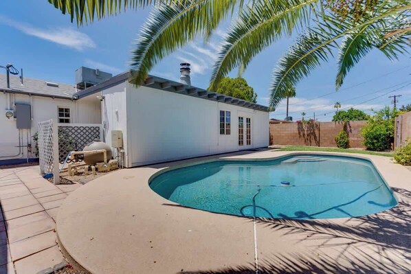 Un jardin à l’arrière d’une maison, avec une piscine, une maison blanche et un mur en briques.