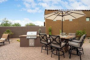 Eine Terrasse im Freien mit Grill, Esstisch, Stühlen und einem großen Sonnenschirm.
