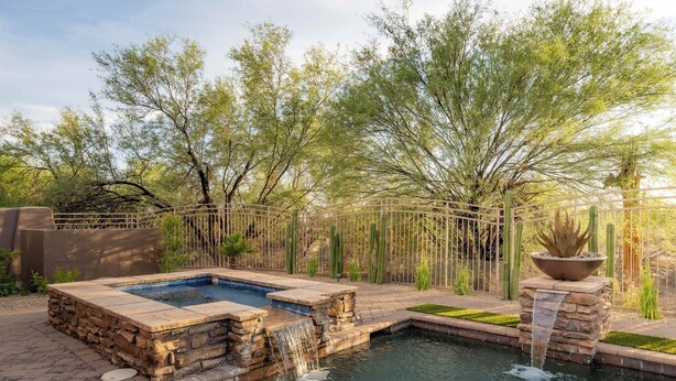 A swimming pool with a waterfall feature, surrounded by a wooden fence and various desert plants. - 4BR The Talon Trail Home: Pool, Spa,  Golf Access (Scottsdale)