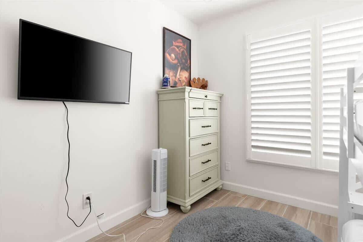 A room with a flat-screen TV mounted on the wall, a white dresser with drawers, a standing fan, and a window with white blinds.