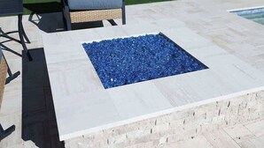 Un área de estar al aire libre con forma cuadrada, un borde blanco y una chimenea de vidrio azul.