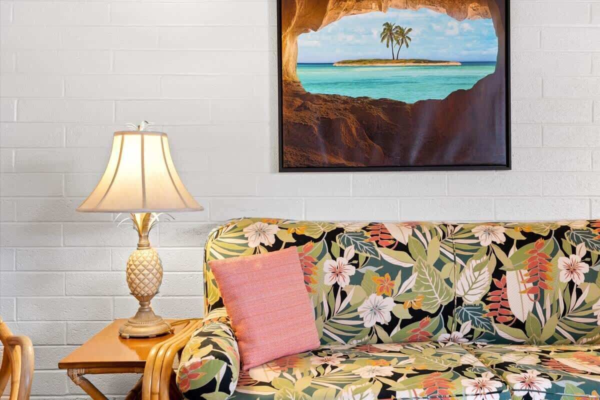 Ein Zimmer mit weißer Ziegelwand, einem Holztischchen mit Lampe, einem blumigen Sofa und einem gerahmten Bild einer tropischen Insel.