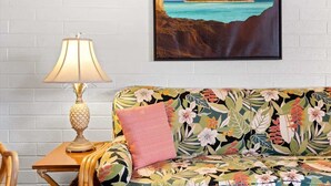 Ein Zimmer mit weißer Ziegelwand, einem Holztischchen mit Lampe, einem blumigen Sofa und einem gerahmten Bild einer tropischen Insel.