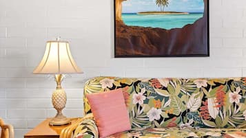 Ein Zimmer mit weißer Ziegelwand, einem Holztischchen mit Lampe, einem blumigen Sofa und einem gerahmten Bild einer tropischen Insel.