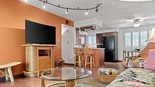 Ein modernes Wohnzimmer mit Flachbildfernseher, Glastisch, Korbmöbeln und einer Küchenzeile im Hintergrund.