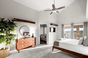 Une chambre à coucher avec un lit, une commode en bois, un ventilateur de plafond, une grande fenêtre et une plante.