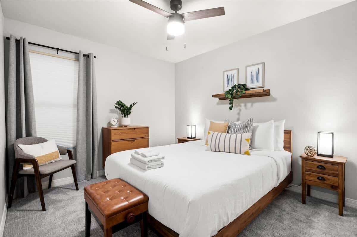 A bedroom with a bed, bedside tables, a chair, a bench, a ceiling fan, and a window with curtains.