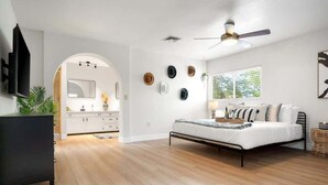 Un dormitorio moderno con cama, ventilador de techo, televisor y una zona de cocina visible a través de un arco.