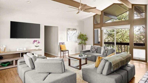 Ein geräumiges Wohnzimmer mit einem großen Flachbildfernseher, grauen Sofas, einem Couchtisch und einem Deckenventilator.