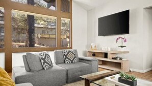 Ein modernes Wohnzimmer mit einem grauen Ecksofa, einem Flachbildfernseher an der Wand und einem großen Fenster mit Blick auf Bäume und ein Gebäude.