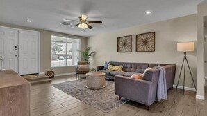 A spacious living room with a grey sofa, a coffee table, and a ceiling fan. There is a large window with blinds, a floor lamp, and a door in the background. - Exclusive retreat- Heated pool & BBQ grill (Peoria)