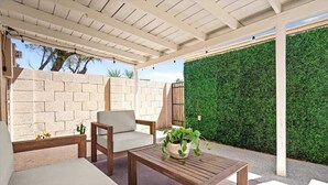 Ein überdachter Patio-Bereich mit Holztisch und gepolstertem Stuhl, einem Blumentopf und einer grünen Heckenwand.