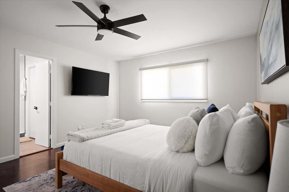 A bedroom with a bed, a ceiling fan, a flat-screen TV, a window, and a door.