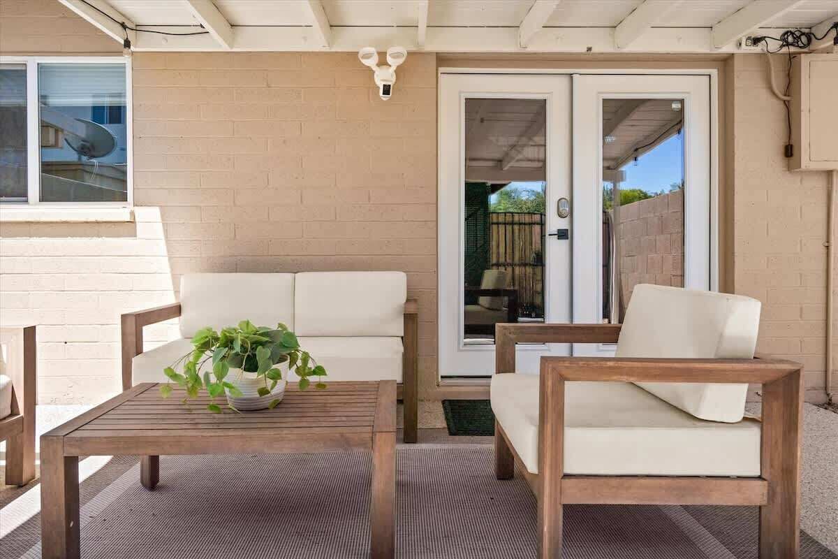 Un espace patio comprenant deux chaises blanches avec coussins, une table en bois ornée d’une plante, et une porte-fenêtre coulissante donnant sur un jardin.