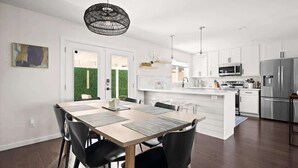 Eine moderne Küche mit weißen Schränken, einer zentralen Kochinsel, Edelstahlgeräten und einem Essbereich mit Holztisch und schwarzen Stühlen.