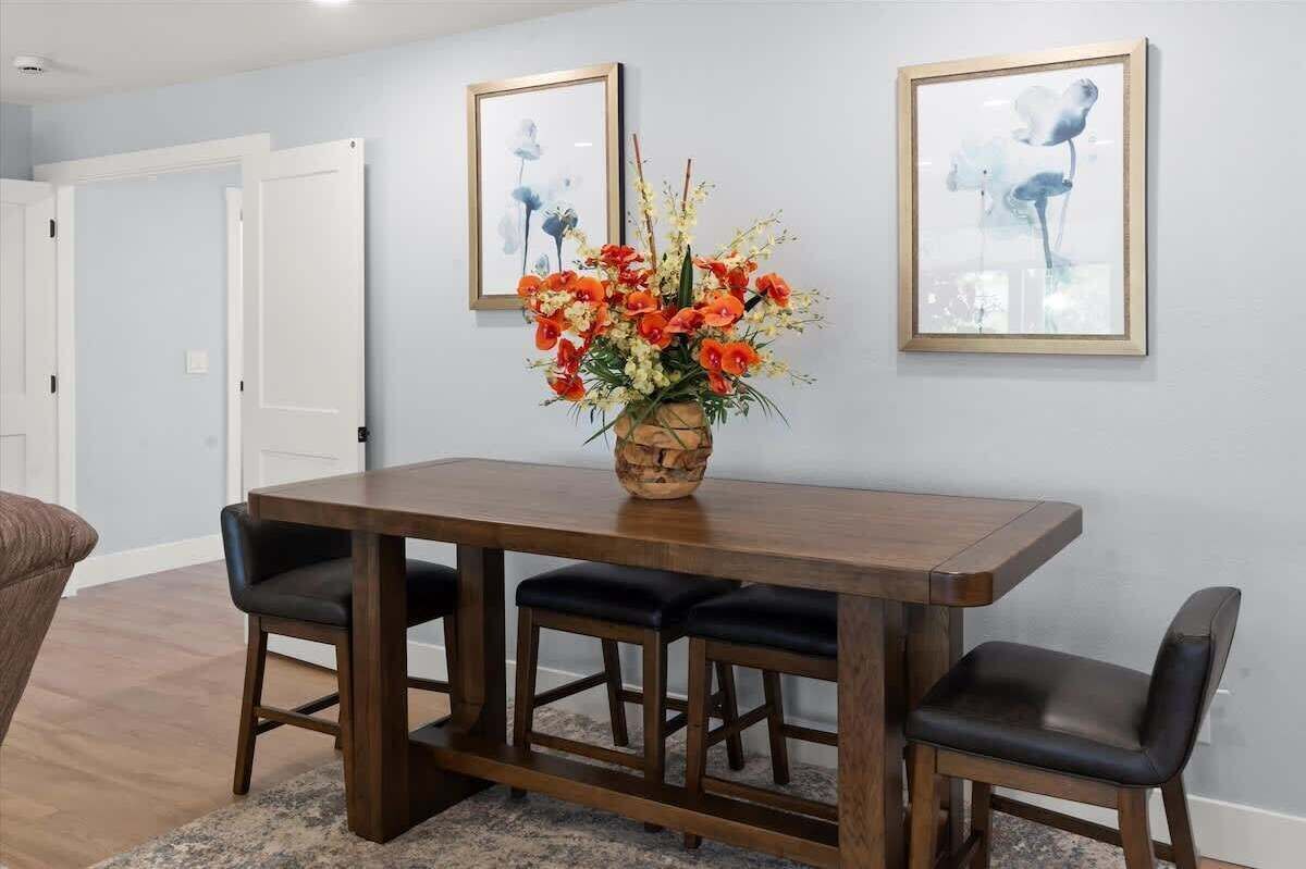 Un comedor con una mesa de madera y sillas tapizadas en negro, un florero con flores y dos cuadros en la pared.