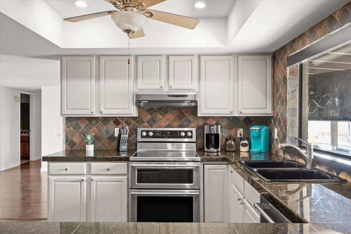 A modern kitchen with white cabinets, stainless steel appliances, a tiled backsplash, and a large window.