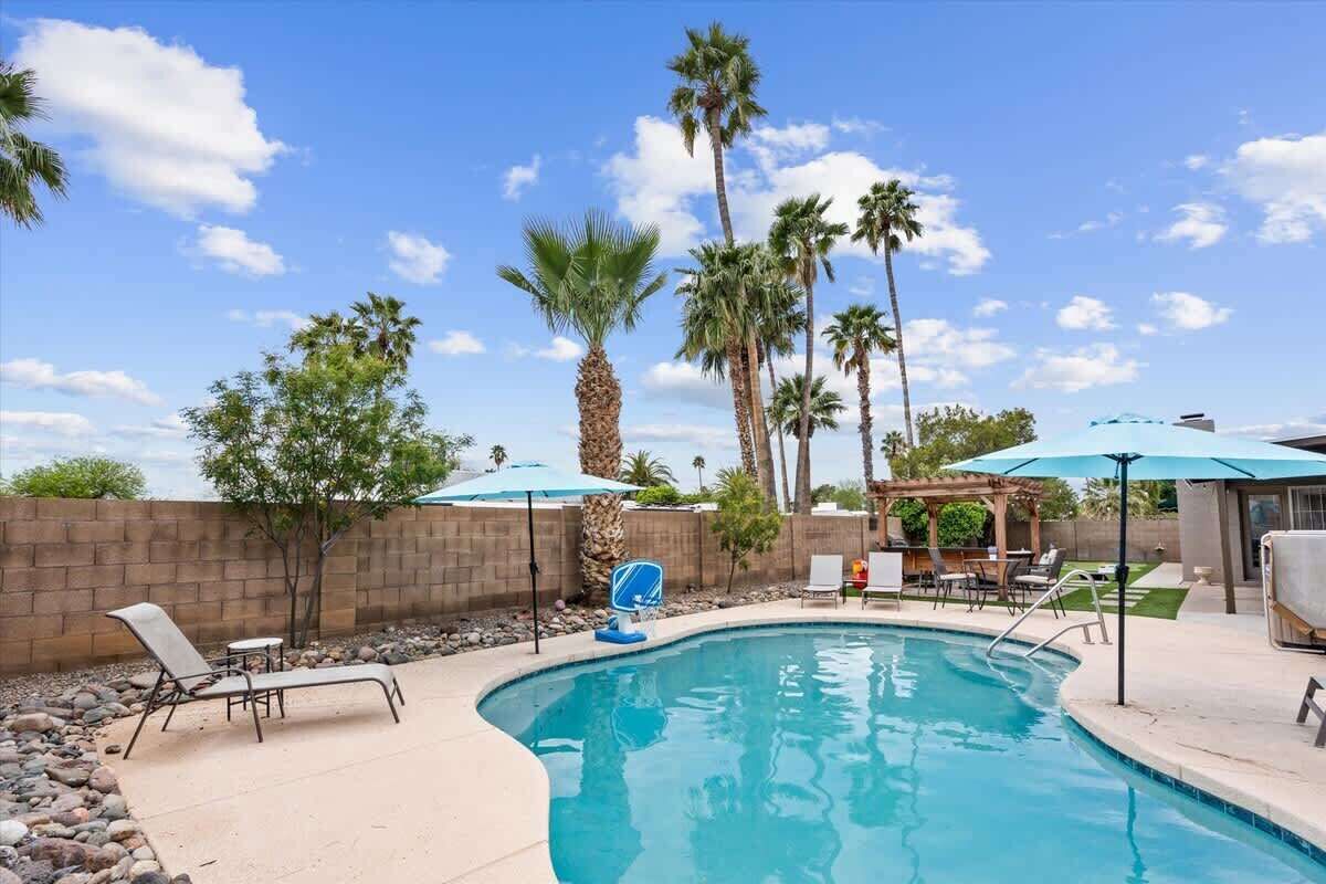 A swimming pool with lounge chairs and umbrellas, surrounded by palm trees and a brick wall.