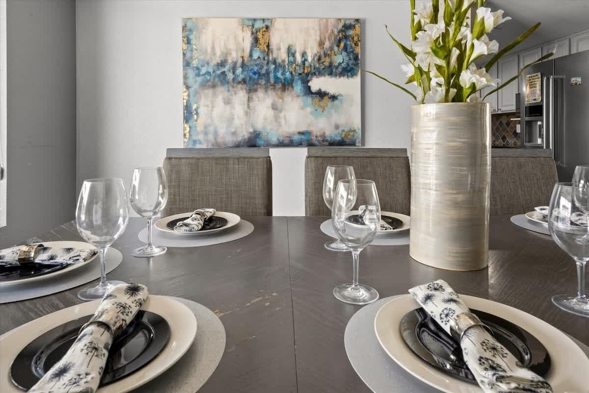 A dining area with a dark wooden table set for four, featuring white plates, black napkins, and clear wine glasses. A large vase with white flowers is on the table, and a modern abstract painting is on the wall behind.