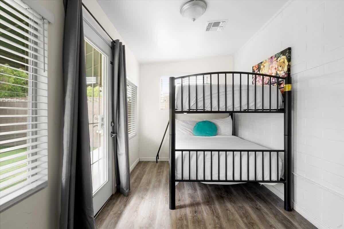 Uma cama beliche em um quarto com porta de vidro deslizante para uma varanda, paredes brancas e piso de madeira.